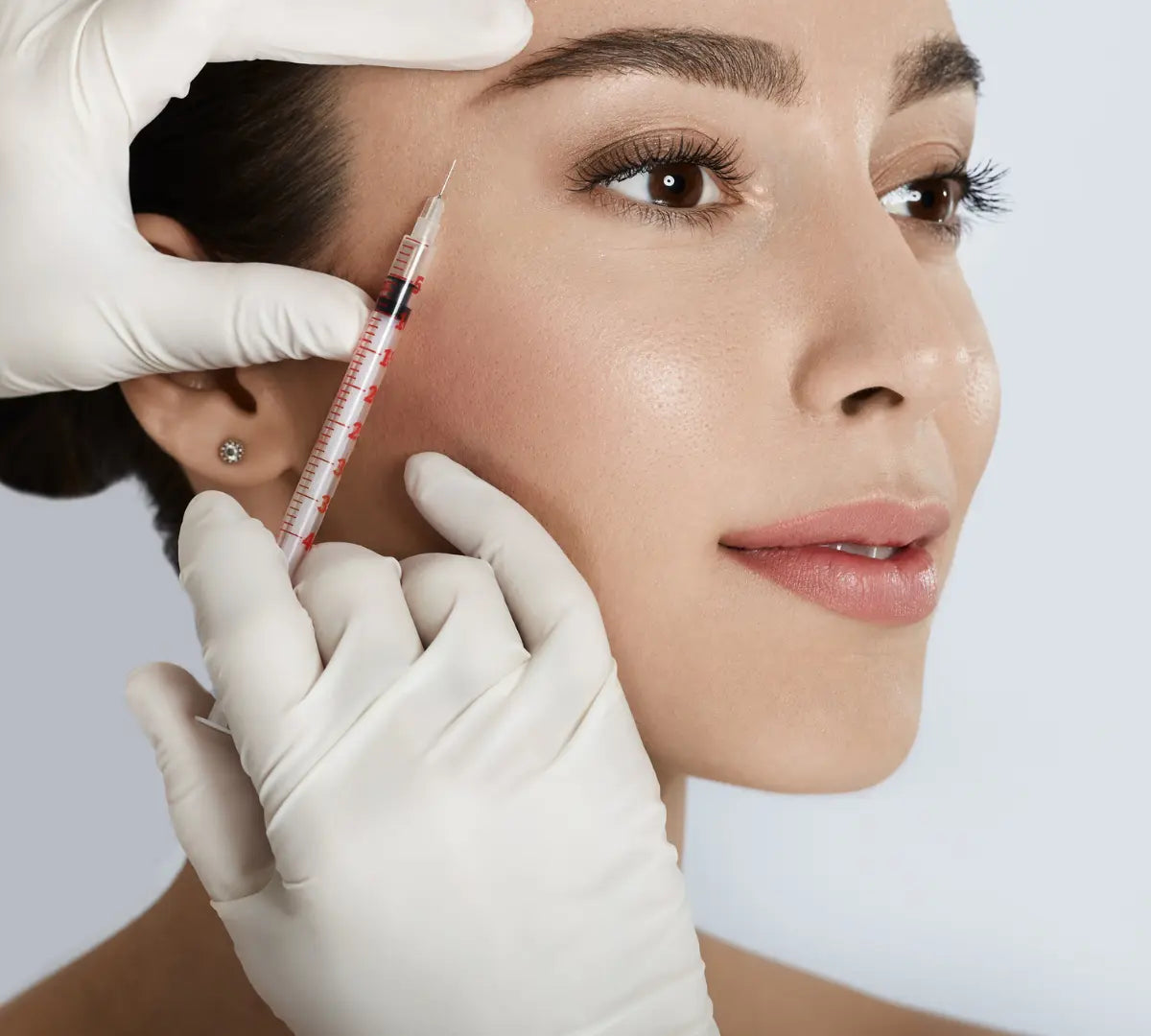 Woman receiving a facial injection with a syringe held by gloved hands against a white background